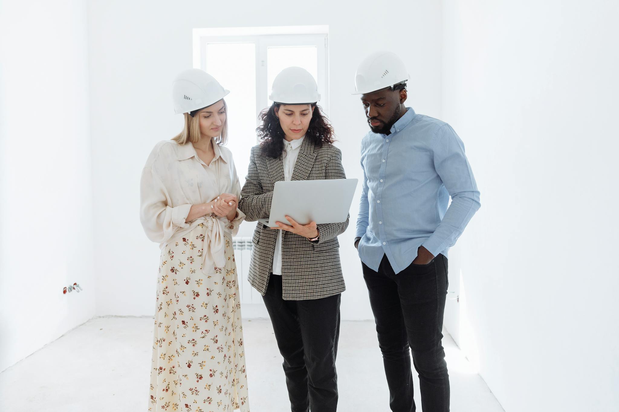 Three professionals in hard hats examining floor plans on a laptop indoors.